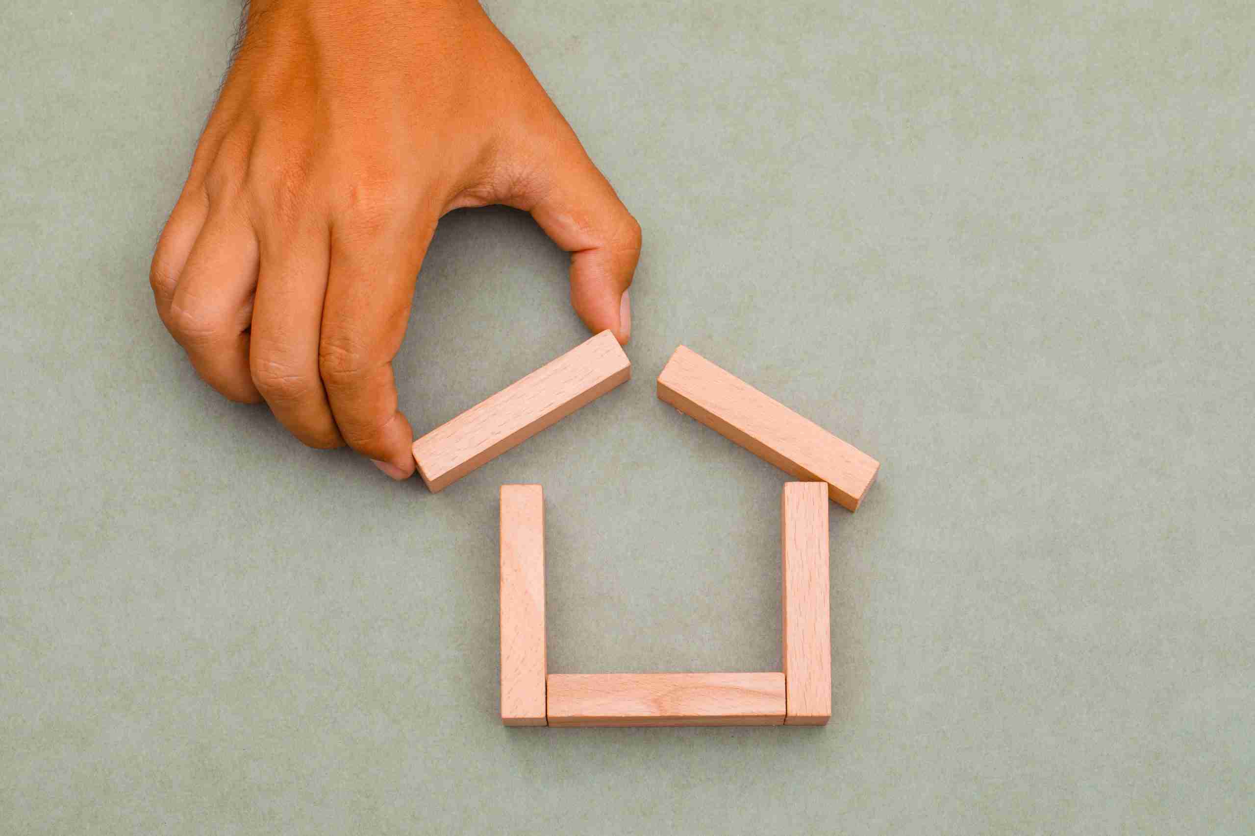 Una mano montando una casa con palos de madera sobre un fondo gris, representando la Nuda propiedad