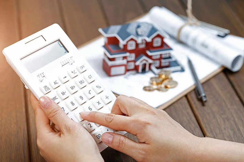 Mano de mujer calcula subrogación de hipoteca, con casa miniatura y documentos de fondo