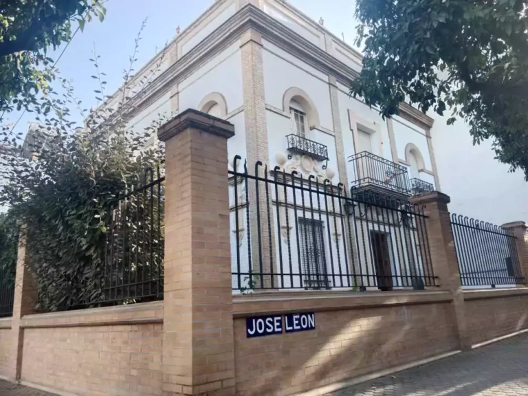 Fotografía exterior de la Calle José León, cuyo nombre se encuentra en un muro de ladrillo y detrás de si hay una casa blanca, típica del barrio de león, sevilla